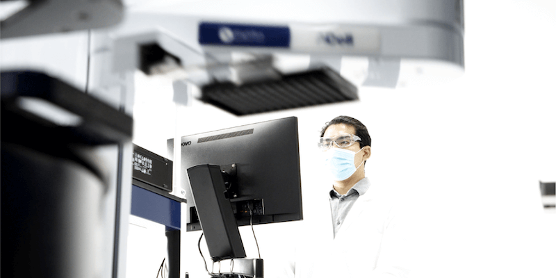 Scientist wearing a lab coat, safety glasses, and a face mask operates an automated laboratory system by HighRes Biosolutions. 